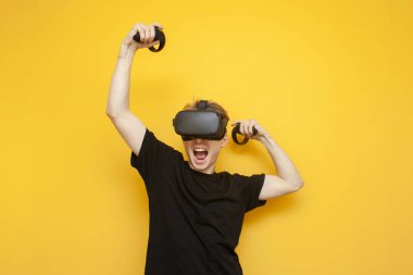 Sarı arka planda VR gözlüklü neşeli bir genç oyun oynuyor ve joysticklerle kazanıyor. Duygusal bir adam sanal bir oyun kazanıyor.