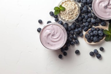 Beyaz bir masada bir bardakta taze yaban mersinli yoğurt, mısır gevreği ve böğürtlen.