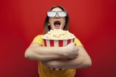 Mutlu genç kız 3D gözlüklü izleyici elinde büyük bir kutu patlamış mısır tutuyor ve kırmızı arka planda bir film izliyor.