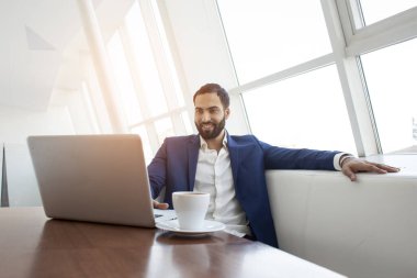 Genç ve başarılı bir işadamı sabah iş yerinde dizüstü bilgisayar kullanır ve kahve içer. Serbest çalışan biri beyaz bir kafede bilgisayarla oturuyor.