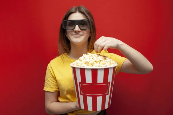 3D gözlüklü güzel bir genç kız patlamış mısır yiyor ve kırmızı arka planda film seyrediyor. — Stok fotoğraf