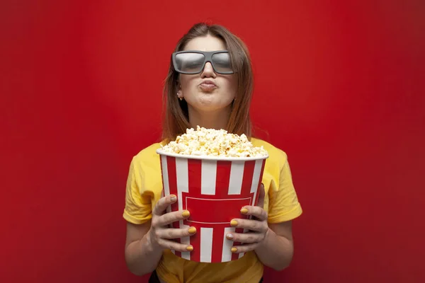 Niña feliz el espectador en gafas 3D sostiene una gran caja de palomitas de maíz y ve una película sobre un fondo de color rojo — Foto de Stock