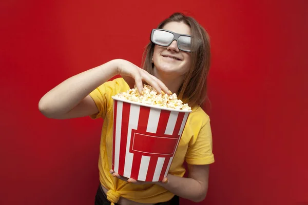 Hermosa joven en gafas 3D comer palomitas de maíz y ver una película sobre un fondo de color rojo — Foto de Stock