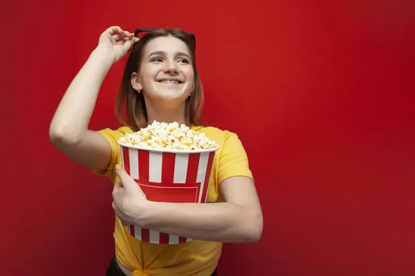 Happy hipster girl with popcorn puts on 3D glasses and smiles on a red colored background — Stock Photo, Image