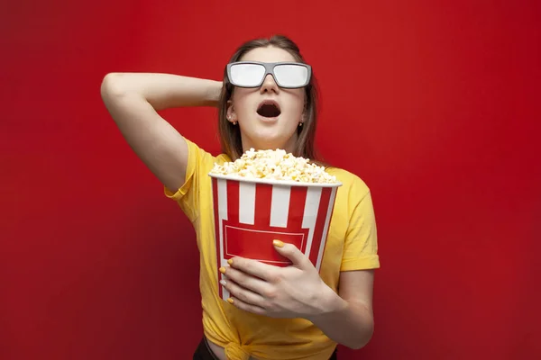 Şaşırmış, şok olmuş bir kız 3D gözlüklü izleyici bir film izliyor ve elinde kırmızı arka planda büyük bir kutu patlamış mısır tutuyor. — Stok fotoğraf