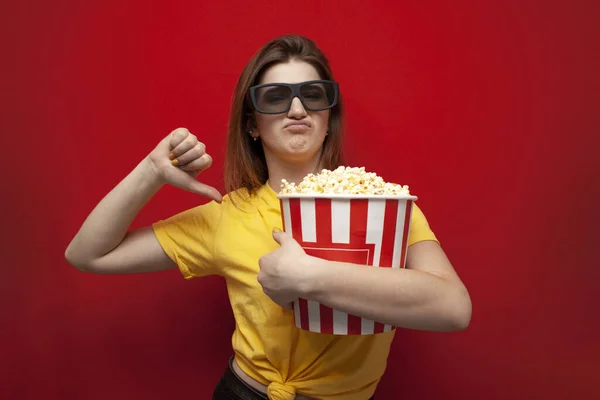 Сумна, незадоволена дівчина в 3D окулярах дивиться поганий фільм з попкорном і показує нелюбов на червоному тлі — стокове фото