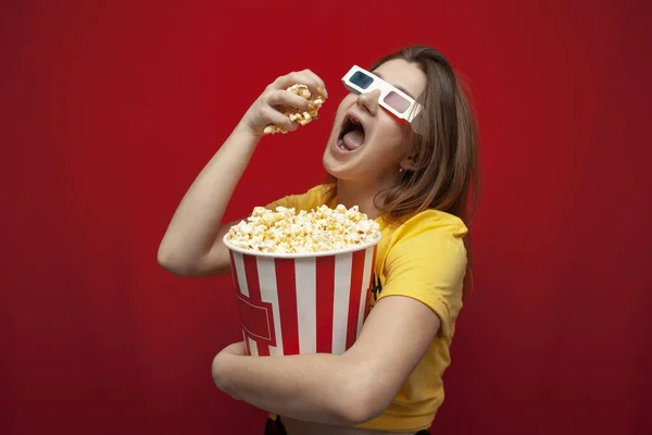 Hermosa joven en gafas 3D comer palomitas de maíz y ver una película sobre un fondo de color rojo — Foto de Stock