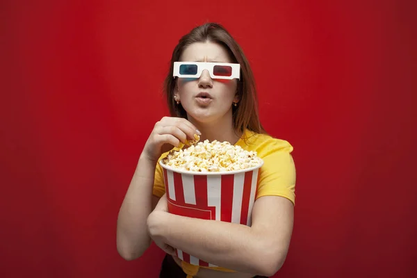 Красива молода емоційна дівчина в 3D окулярах їсть попкорн, дивиться фільм і кричить на червоному тлі, здивований глядач — стокове фото