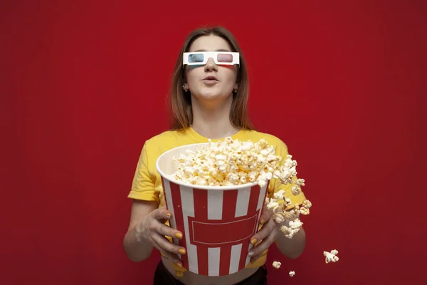Divertida joven sorprendida en gafas 3D viendo una película y comiendo palomitas de maíz sobre un fondo de color rojo, ella grita y palomitas de maíz vuela — Foto de Stock