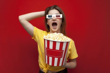 funny happy young girl in 3D glasses and with popcorn on a red background, a student watching a 3D film, copy space