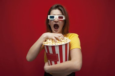 funny young girl student watching a movie in 3D glasses and screaming on a red background, she eats popcorn and is delighted