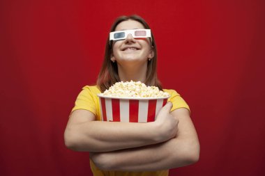 funny happy young girl in 3D glasses and with popcorn on a red background, a student watching a 3D film