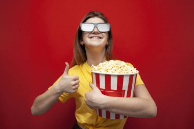 Üç boyutlu gözlüklü neşeli genç kız, patlamış mısırlı bir film izliyor ve kırmızı bir arka planda gibi görünüyor.