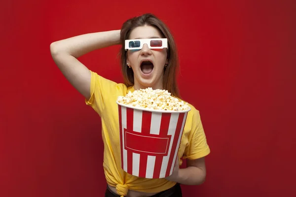 Funny happy young girl in 3D glasses and with popcorn on a red background, a student watching a 3D film, copy space — Stock Photo, Image