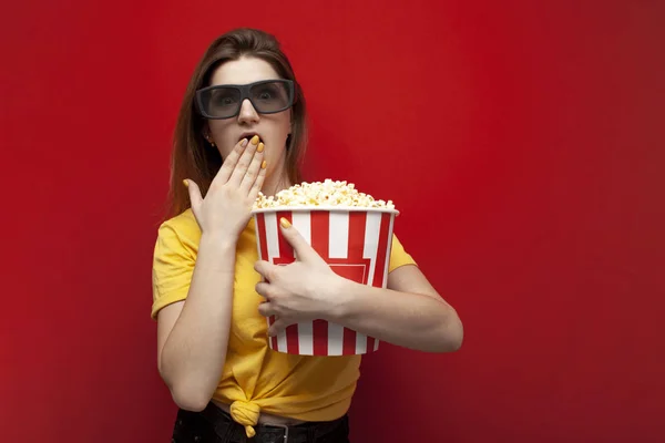 Surprised young girl student watching a movie in 3D glasses and eating popcorn on a red background — ストック写真