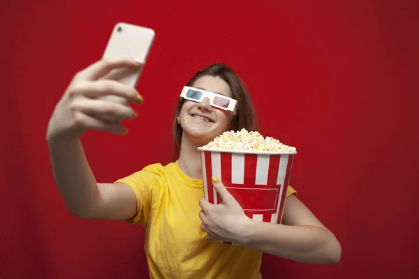 Attractive girl in 3D glasses and with popcorn is watching a movie on a smartphone, taking a selfie on a red background — Stok fotoğraf