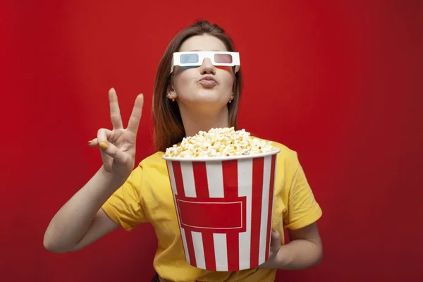 Divertida joven feliz en gafas 3D y con palomitas de maíz sobre un fondo rojo, un estudiante viendo una película 3D — Foto de Stock