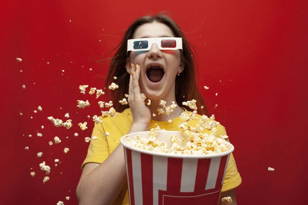 Divertida joven sorprendida en gafas 3D viendo una película y comiendo palomitas de maíz sobre un fondo de color rojo, ella grita y palomitas de maíz vuela — Foto de Stock