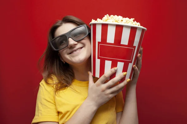 Mutlu genç kız 3D gözlüklü izleyici elinde büyük bir kutu patlamış mısır tutuyor ve kırmızı arka planda bir film izliyor. — Stok fotoğraf