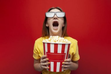 Mutlu genç kız 3D gözlüklü izleyici elinde büyük bir kutu patlamış mısır tutuyor ve kırmızı arka planda bir film izliyor.