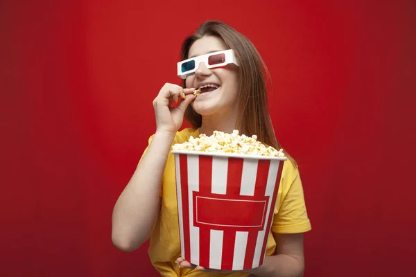 Beautiful young girl in 3D glasses eating popcorn and watching a movie on a red color background — Stock Photo, Image