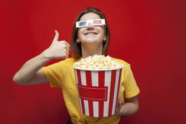 Üç boyutlu gözlüklü neşeli genç kız, patlamış mısırlı bir film izliyor ve kırmızı bir arka planda gibi görünüyor.