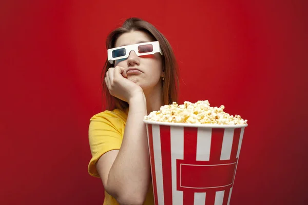 Triste joven viendo una película mala y aburrida en gafas 3D y con palomitas de maíz sobre un fondo rojo — Foto de Stock