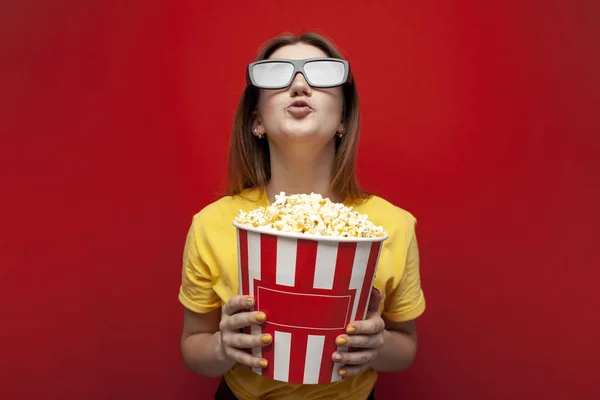Happy young girl the viewer in 3D glasses holds a big box of popcorn and watches a movie on a red color background — Stock Photo, Image