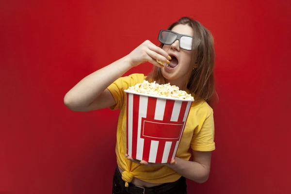 Hermosa joven en gafas 3D comer palomitas de maíz y ver una película sobre un fondo de color rojo — Foto de Stock