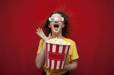 funny young girl student watching a movie in 3D glasses and screaming on a red background, she eats popcorn and is delighted, copy space