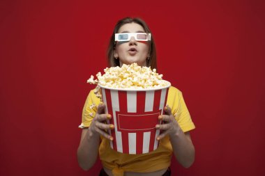 surprised young girl student watching a movie in 3D glasses and eating popcorn on a red background, copy space