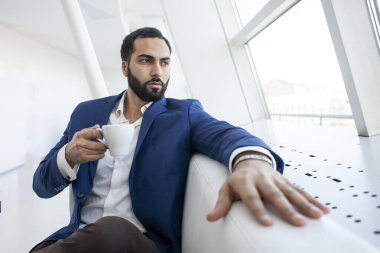 Takım elbiseli başarılı bir iş adamı bir kafede oturur, kahve içer ve pencereden dışarı bakar.