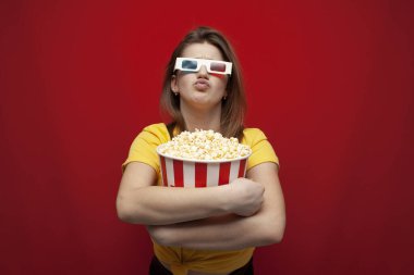 surprised young girl student watching a movie in 3D glasses and eating popcorn on a red background, copy space