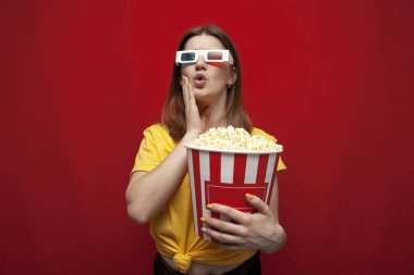 surprised young girl student watching a movie in 3D glasses and eating popcorn on a red background, copy space