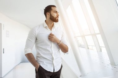Portrait of a successful handsome man in a white shirt in the morning in a white building, businessman looks out the window, copy space