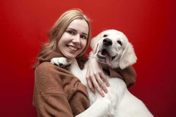 happy cheerful beautiful girl holding a dog on a red background, a woman hugs a golden retriever puppy and smiles, people with pets, copy space