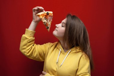 Genç mutlu kız, renkli bir arka planda lezzetli bir dilim pizzayı kokluyor. Lezzetli bir fast food kokusu.