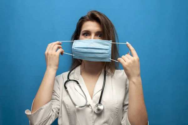 Ung flicka sjuksköterska sätter på en virusmask på en blå bakgrund, skydd av coronavirus epidemin — Stockfoto