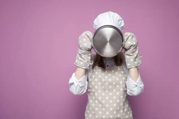 부엌에서 요리하는 소녀가 분홍색 배경에 냄비로 얼굴을 가리고 부엌에서 물건을 — 스톡 사진
