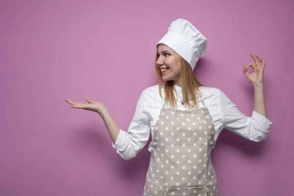快乐的女孩穿着围裙做饭 面带微笑地看着一个地方 那里有彩色背景的文字 快乐的女人穿着厨房的衣服 复制空间 — 图库照片