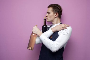 Profesyonel, genç ve yakışıklı garson pembe arka planda iki şişe kırmızı ve beyaz şaraba sarılıyor, alkol aşkı kavramı, fotokopi alanı