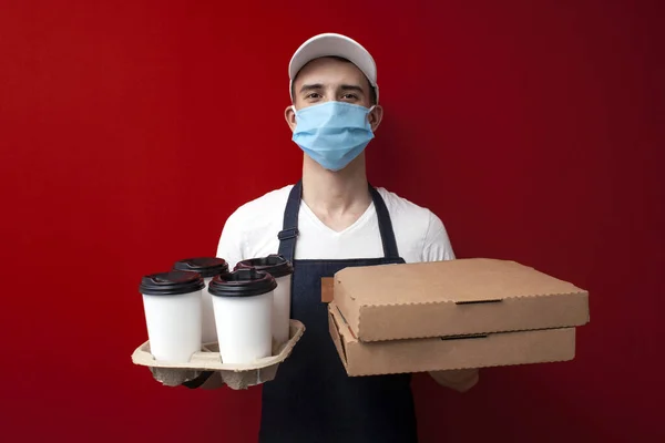 Korunaklı maskeli ve eldivenli genç profesyonel yemek dağıtıcısı pizza ve kahve kutularını tutuyor ve Coronavirus salgını sırasında müşteriye hızlı doğum yaptırıyor.