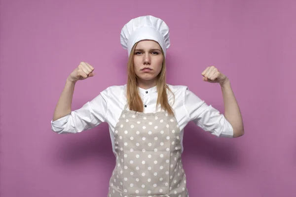 穿着围裙的年轻而严肃的强壮姑娘在粉红的背景上显得很有力量 穿着厨房衣服的家庭主妇也很有力量 — 图库照片