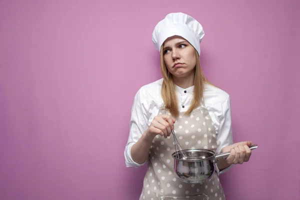 悲伤的女孩穿着围裙做饭 拿着碗碟 用粉红的背景混合食物 疲惫的家庭主妇拿着厨房用具 看着一处文字的地方 复制空间 — 图库照片