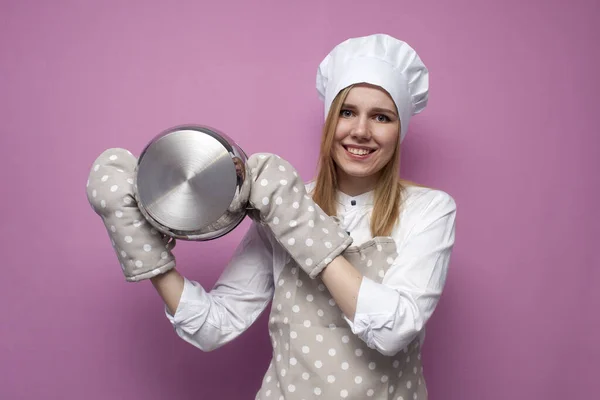 穿着厨房衣服快乐漂亮的女厨师戴着粉色背景的手套拿着平底锅 女家庭主妇拿着厨房用具 复制空间 — 图库照片