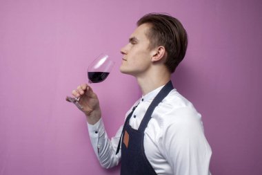 Genç şarap garsonu, arka planda kırmızı şarap kokusunu kontrol eder. Beyaz gömlekli bir şarap uzmanı ve önlüklü bir adam bardağı tutar. Garson şarabın tadına bakar.