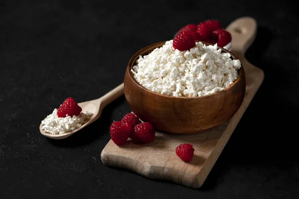 fresh homemade pure cottage cheese in a wooden plate with raspberries in a rustic bowl against a dark background, healthy food on a black table, copy space