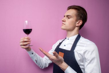 Üniformalı genç profesyonel garson bir bardak kırmızı şarap tutuyor ve elini pembe arka planda gösteriyor. Bir şarap garsonu şarap öneriyor.
