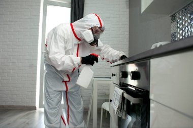 Koruyucu giysili ve solunum cihazlı bir adam mobilyaları kimyasal bir maddeyle siliyor, odayı dezenfekte ediyor, hijyenik işler yapıyor.