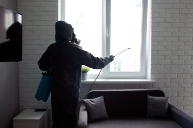 Evde dezenfeksiyon. Koruyucu giysili bir adam, evi koronavirüsten koruyan kimyasal bir maddeyle pencereyi temizliyor.